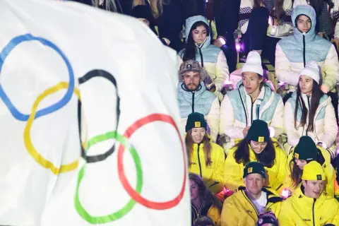 Młodzi sportowcy w barwach narodowych, z flagą olimpijską w tle, czekają na start reprezentacji Polski na Zimowe Igrzyska Olimpijskie 2026.