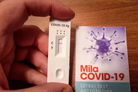 Ręka trzyma test antygenowy COVID-19 z jedną kreską. Obok opakowanie z grafiką wirusa i napisem "Szybki test antygenowy".