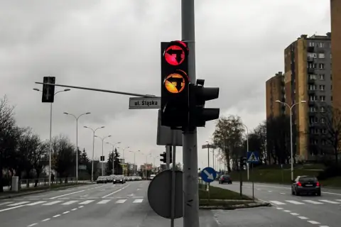 Sygnalizator S3 z czerwonym i żółtym światłem kierunkowym w lewo, na tle pochmurnego nieba i bloków mieszkalnych.