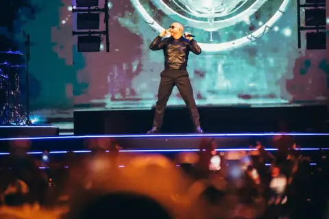 Pitbull Polska na scenie, w czarnej kurtce i spodniach, śpiewa do mikrofonu. Tłum w dole nagrywa koncert.