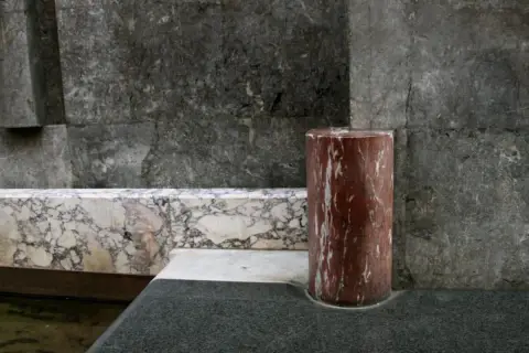 Cylindryczny pomnik z konglomeratu w odcieniach czerwieni i bieli, na tle szarej, kamiennej ściany i marmurowej ławy.