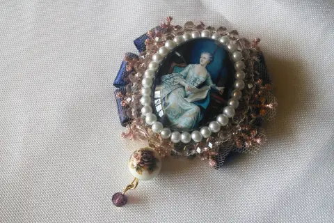 Broche con camafeos, perlas y detalles florales. Una dama elegante lee un libro en un retrato ovalado, rodeada de encaje y cristales.