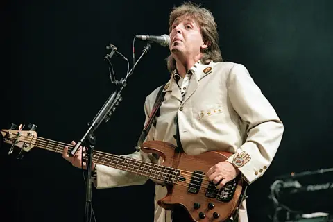 Paul McCartney z zespołu The Beatles gra na basie podczas koncertu, ubrany w elegancki beżowy garnitur.