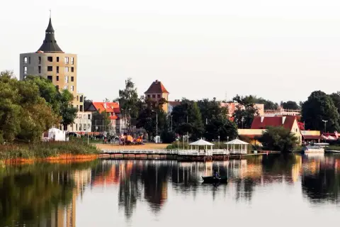 Widok na jezioro z łodzią, pomostem z altanami i budynkami w tle.
