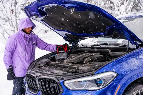 Mężczyzna w fioletowej kurtce podłącza booster do akumulatora niebieskiego BMW. To rozwiązanie, co może pobierać prąd w samochodzie na postoju, gdy jest zimno.