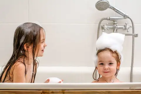 Dwie dziewczynki w wannie, jedna z pianą na głowie, druga z mokrymi włosami. Radosne mycie głowy dziecku to świetna zabawa.