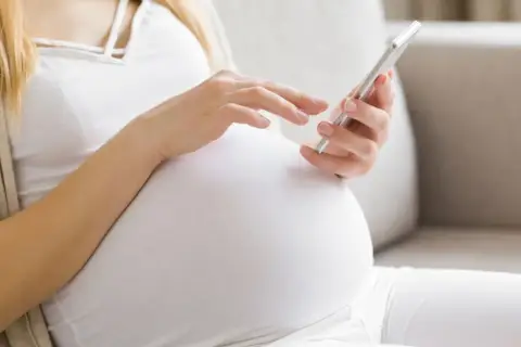 Kobieta w 24. tygodniu ciąży (l4 w ciąży) przegląda coś na telefonie, siedząc na kanapie.