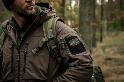 Mężczyzna w brązowej kurtce z naszywką Helikon-Tex, symbol polskie marki sportowe, w lesie.