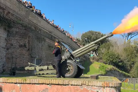 Il cannone Gianicolo spara una palla di fumo arancione e gialla. Un soldato in uniforme osserva, mentre una folla guarda dall'alto.
