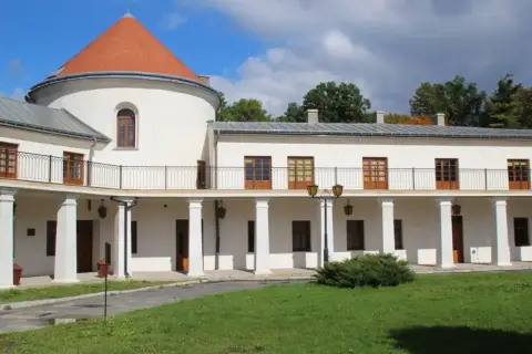 Zamek w Lesku, biały budynek z okrągłą wieżą i pomarańczowym dachem, otoczony zielenią.