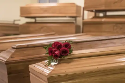 Wiele rodzajów trumien, niektóre ułożone piętrowo. Na jednej z nich bukiet czerwonych róż i złota obręcz.