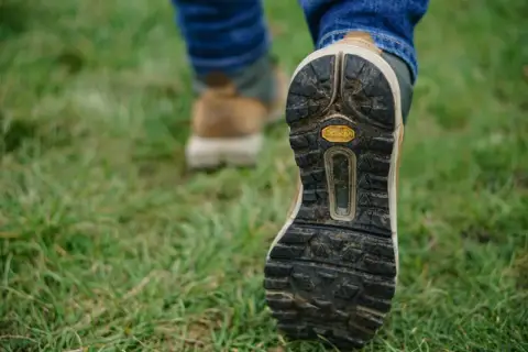 Osoba w dżinsach i butach trekkingowych stawia kroki na trawie. Czy buty trekkingowe powinny być większe? Zobacz podeszwę Vibram.