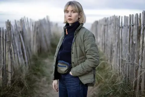 Karen Pirie står vid en vindpinad strand, klädd i en grön jacka och jeans. Hon bär en midjeväska, redo för ett nytt fall.