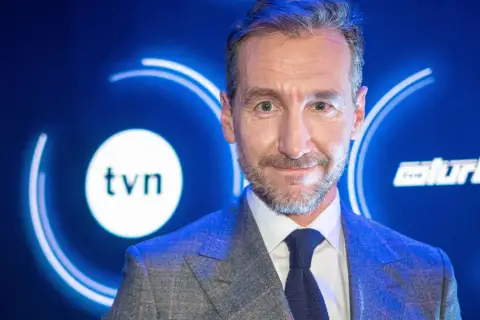 Uśmiechnięty mężczyzna w garniturze na tle logo TVN. Jeden z dziennikarzy Wiadomości TVN, gotowy do przekazania najnowszych informacji.