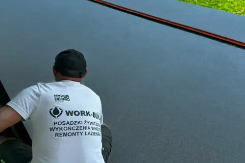 Pracownik w koszulce z napisem "Posadzki żywiczne" wykonuje prace na ciemnej, gładkiej powierzchni.