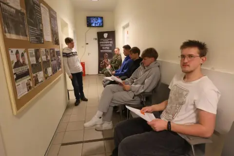 Młodzi ludzie czekają na komisję wojskową, przeglądając dokumenty. Na ścianie wiszą plakaty zachęcające do wstąpienia do wojska.
