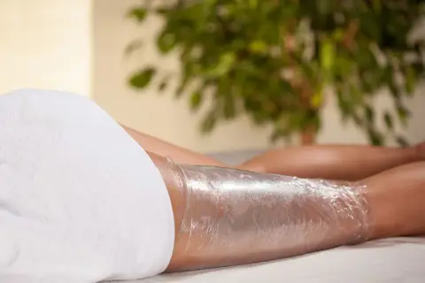 Zabieg body wrapping z użyciem kosmetyków. Nogi owinięte folią, przygotowane na odżywcze działanie.