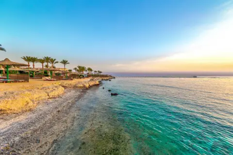 Piękna pogoda w Marsa Alam: piaszczysta plaża z palmami, leżakami i parasolami, turkusowe morze i długie molo na horyzoncie.