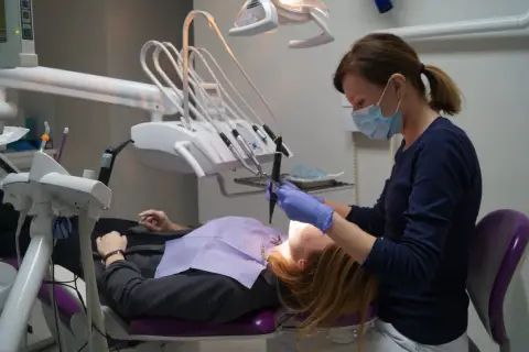 Dentystka w masce i rękawiczkach pracuje przy pacjencie. W gabinecie widoczny sprzęt do stomatologii.