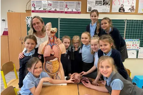 Nauczycielka i uczennice przy modelu anatomicznym. Jedna z dziewczynek trzyma serce na dłoni, jakby ofiarowywała je w geście nauki.