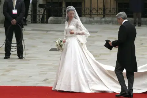 Kate Middleton w drugiej sukni ślubnej, z welonem i bukietem, idzie po czerwonym dywanie. Obok niej mężczyzna w żakiecie trzyma cylinder.