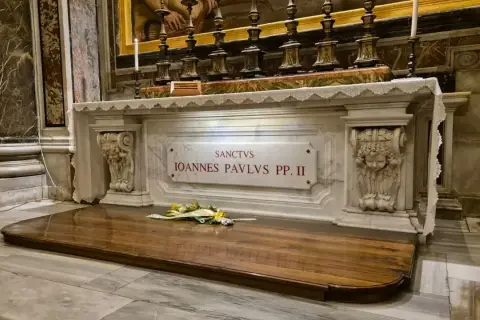 Grób świętego Jana Pawła II w szklanej trumnie, ozdobiony kwiatami.