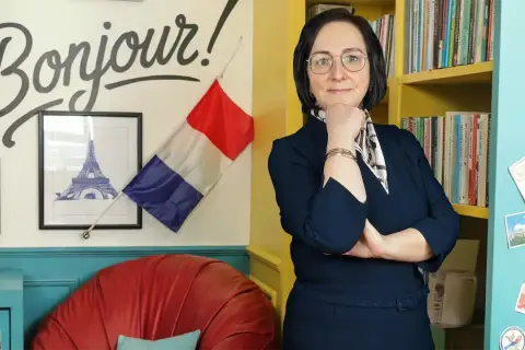 Kobieta w okularach stoi przed ścianą z napisem "Bonjour!", francuską flagą i wieżą Eiffla.