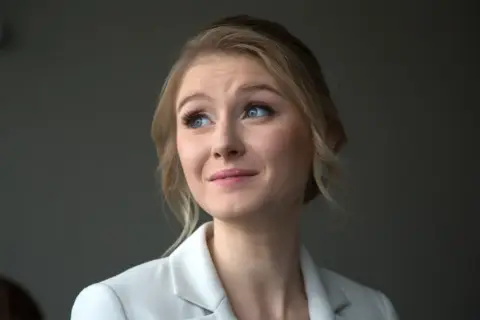 Portret młodej kobiety z blond włosami i niebieskimi oczami, w białej marynarce. Jej wiek to około 30 lat.