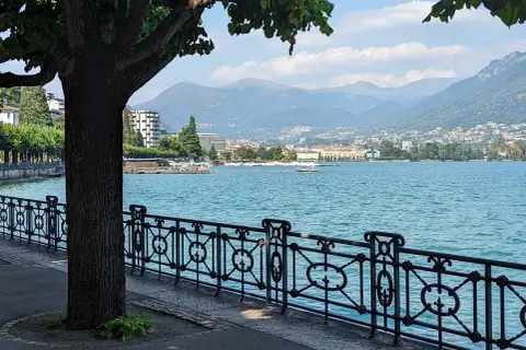 Vue paisible du lac de Lugano, avec une promenade bordée d'arbres et de montagnes en arrière-plan.