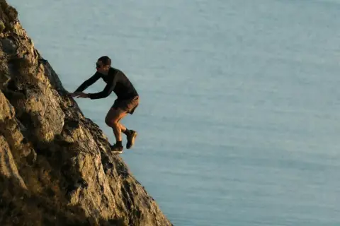 Kilian Jornet escalade une paroi rocheuse abrupte, dominant un plan d'eau calme, rappelant l'esprit d'aventure dans les Pyrénées.