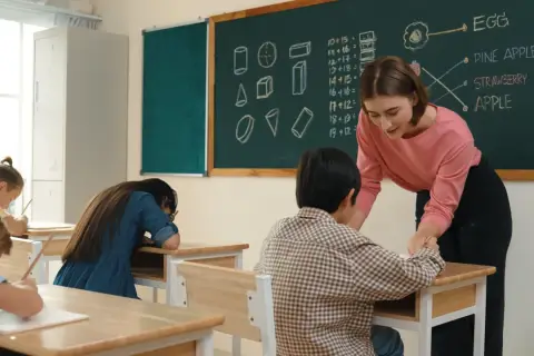 Nauczycielka pomaga uczniowi przy biurku, gdy jest wystawienie ocen. Na tablicy matematyka i owoce.