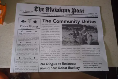 Gazeta "The Hawkins Post" z artykułem "The Community Unites" i zdjęciem dzieci sprzątających plażę.