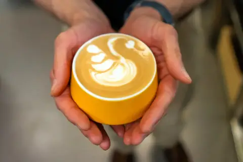 Dłonie trzymają żółtą filiżankę z kawą, na której artystycznie uformowano łabędzia. Zastanawiasz się, ile kofeiny ma espresso z ekspresu?
