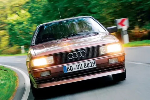 Klasyczny, brązowy Audi quattro z lat 80. pędzi po krętej, leśnej drodze.