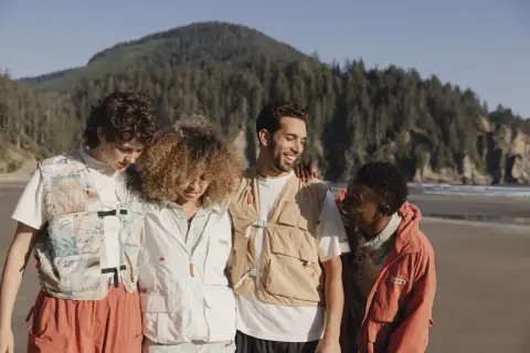 Grupo de amigos con estilo gorpcore en la playa, con chalecos utilitarios y chaquetas, disfrutando del paisaje costero.