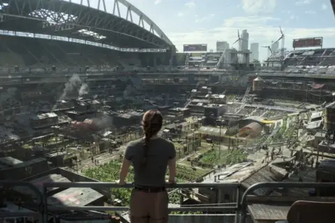 Widok na postapokaliptyczne miasto wewnątrz stadionu, gdzie ludzie żyją w prowizorycznych domach. Może to być scena z The Last of Us sezon 3.