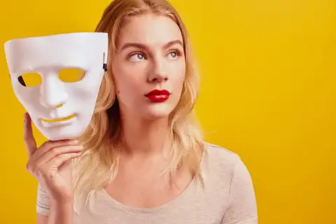 Blondynka z czerwoną szminką trzyma białą maskę, zastanawiając się nad hipokryzją.