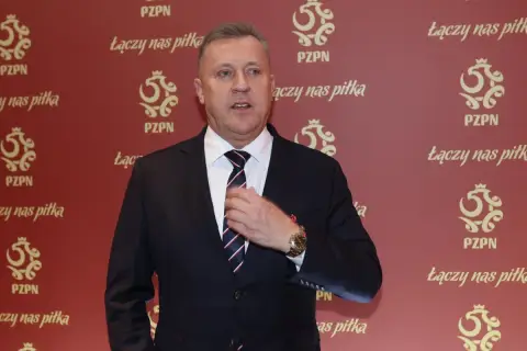 Prezes PZPN w garniturze, poprawiający krawat. Tło ozdobione logotypami PZPN i hasłem "Łączy nas piłka".