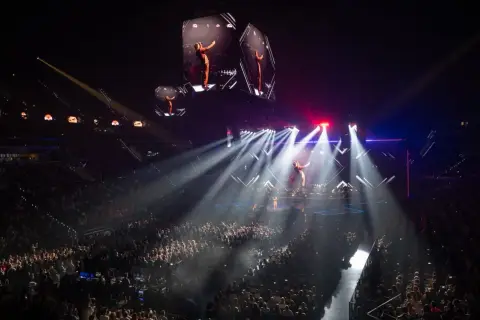 Un artista en un escenario iluminado por focos, con pantallas gigantes mostrando su imagen. ¡Imagina los conciertos Valencia 2026!