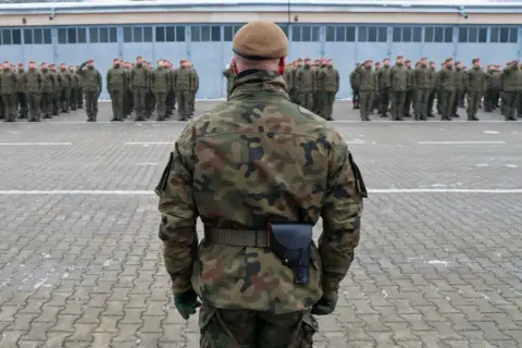 Żołnierze w mundurach polowych stoją na placu apelowym. W tle budynek koszar. To może być początek obowiązkowej służby wojskowej.
