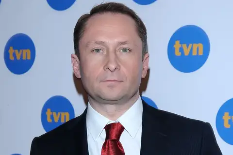 Kamil Durczok w garniturze i czerwonym krawacie na tle logo TVN. Prowadzący Fakty TVN.