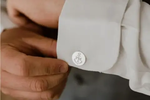 Mężczyzna zapina spinki do mankietów koszuli. Na spinkach widnieje kotwica i inicjały PM. To prosty sposób, jak zapiąć spinki do koszuli.