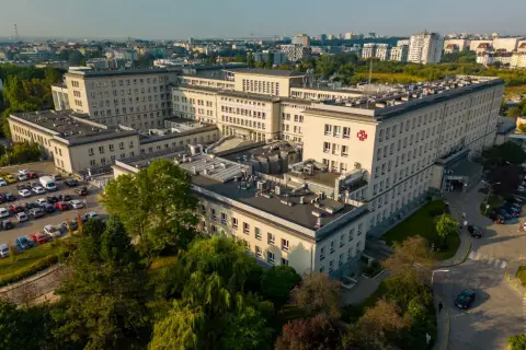Widok z lotu ptaka na duży kompleks szpitalny z czerwonym krzyżem na fasadzie, otoczony drzewami i parkingiem.