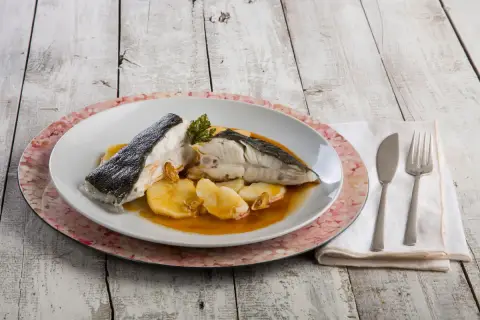 Rodaballo gallego servido con patatas y salsa, acompañado de cubiertos y servilleta sobre una mesa de madera.
