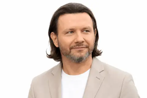 Mężczyzna z siwiejącą brodą i długimi włosami, ubrany w jasną marynarkę, patrzy w bok. Jego **majdan wzrost** jest imponujący.