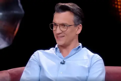 Michał Czernecki w jasnej koszuli i okularach, uśmiecha się. W tle widać fragmenty scenografii, przypominające o jego filmach, serialach i programach.