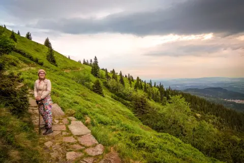 Kobieta z kijkami na kamiennym szlaku, podziwia widok na zielone zbocze i las. Widać, ile się wchodzi na Śnieżkę.