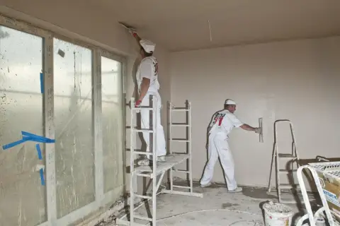 Dwóch fachowców w białych kombinezonach, używając kielni, wyrównuje ściany gładzią.