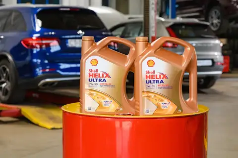 Dwa kanistry oleju silnikowego Shell Helix Ultra 0W-30 stoją na czerwonym bębnie. W tle widać samochody w warsztacie.