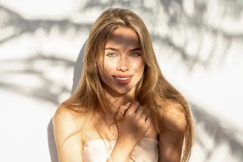 Strahlendes Sommer Make-up: Eine junge Frau mit langen blonden Haaren und grünen Augen lächelt in die Kamera, während Sonnenlicht durch Schatten fällt.